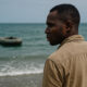 African man gazing at the sea with an abandoned inflatable boat in the distance, symbolizing the impact of visa denials and dangerous migration routes.