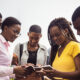 A group of diverse, young African professionals collaborates, using a smartphone and notebook, symbolizing Africa's youthful population driving innovation, technology, and economic growth.