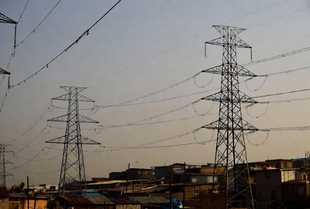 High-voltage power lines stretching over informal settlements in Africa and the Caribbean, where homes below remain without electricity, illustrating energy inequality and lack of grid access despite nearby infrastructure