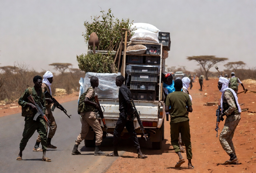 Armed militants on a dusty Sahel road blocking a humanitarian vehicle carrying tree-planting supplies and Ebola response equipment, symbolizing how armed groups obstruct climate and health projects in Africa.