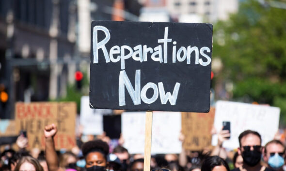 Black Lives Matter protest sign reading 'Reparations Now' with blurred crowd in background, symbolizing ongoing debate over slavery compensation.