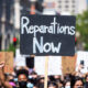 Black Lives Matter protest sign reading 'Reparations Now' with blurred crowd in background, symbolizing ongoing debate over slavery compensation.