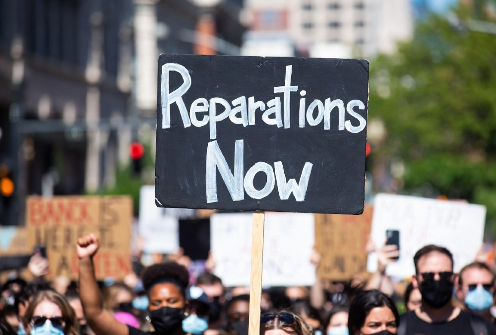Black Lives Matter protest sign reading 'Reparations Now' with blurred crowd in background, symbolizing ongoing debate over slavery compensation.
